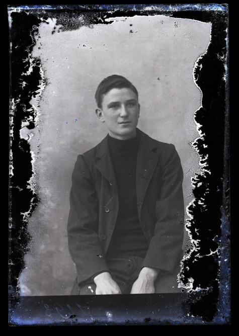 Zittend portret van jongeman in kostuum met halsdichte pullover, kuifvormig gekamd haar met linkerscheiding, Melle , 1910-1920