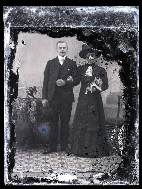 Staand portret van een koppel in feestkledij, man in kostuum met wit hemd en witte vlinderdas en strak gekamd haar, bolhoed in de hand, vrouw met hoed en boementuiltje in de linkerhand , Melle , 1910-1920