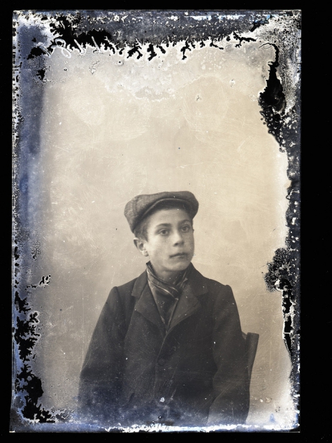 Zittend portret van jonge man in feestkledij met vest en sjaal om de hals, pet op het hoofd, Melle, 1910-1920