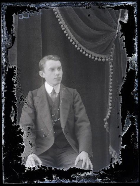 Zittend portret van jonge man in feestkledij met kostuum en witte hemdskraag met stropdas, kuifvormig naar links gekamd haar, Melle, 1910-1920