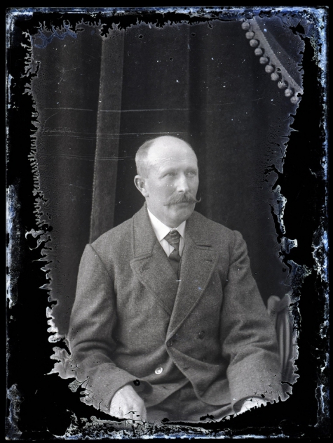 Zittend portret van man in feestkledij met witte hemdsboord en donkere stropdas, gemodelleerde snor en kalend voorhoofd, Melle, 1910-1920