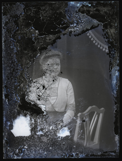 Zittend portret van jonge vrouw in feestkledij bestaande uit donkere rok en bleek vestje met driekwart-mouwen, opgestoken haar, Melle, 1910-1920