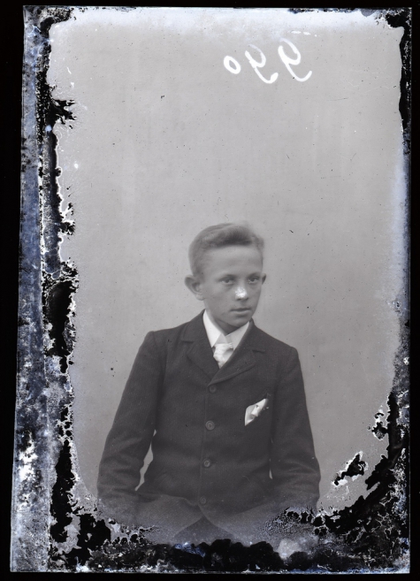 Zittend portret van jongeman in feestkledij bestaande uit kostuum, wit hemd met stropdas en pochdoekje in linker bovenzakje, kuifvormig naar rechts gekamd haar , Melle, 1910-1920
