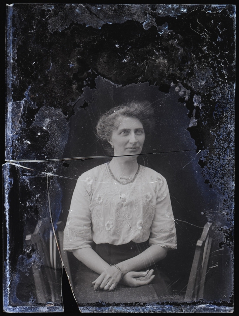 Zittend portret van jonge vrouw in feestkledij bestaande uit lange rok en geborduurde bloes, armband en halsketting, opgestoken haar , Melle, 1910-1920