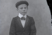 Zittend portret van jongen in kostuum met wit hemd en vlinderdas, pet als hoofddeksel, Melle , 1910-1920