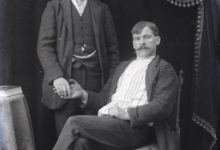 Portret van staande man in feestkledij met wit hemd en stropdas en zittende man, beiden met snor en kuifvormig gekamd haar , Melle , 1910-1920
