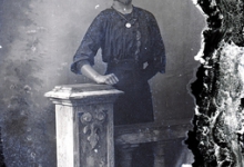 Staand portret van jonge vrouw in feestkledij bestaande uit lange rok en bloes met driekwart mouwen en witte halskraag, halsketting met muntstuk, Melle, 1910-1920