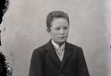 Zittend portret van jongen in feestkledij bestaande uit kostuum,wit hemd en stropdas, golvend naar achter gekamd haar, Melle, 1910-1920