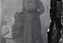 Staand portret van jonge vrouw in feestkledij bestaande uit lang kleed met brede heupband en driekwart mouwen, opgestoken haar, Melle, 1910-1920