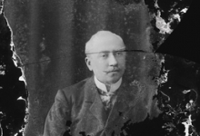 Zittend portret van man in feestkledij bestaande uit kostuum en wit hemd met stropdas, gemodelleerde snor, Melle, 1910-1920